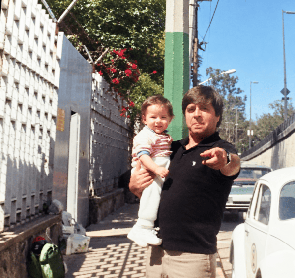 Un padre sostiene a su hijo de aproximadamente un año y señala hacia la cámara en una calle soleada.